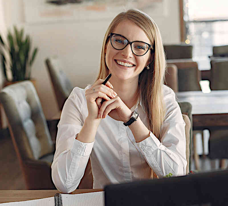 Frau mit Brille und Stift in der Hand sitzt lächelnd im Büro | Symbolbild