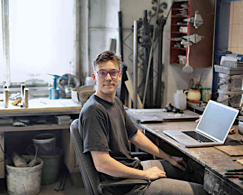 Junger Mann sitzt in einer Tischler-Werkstatt am Schreibtisch vor einem Laptop | Symbolbild
